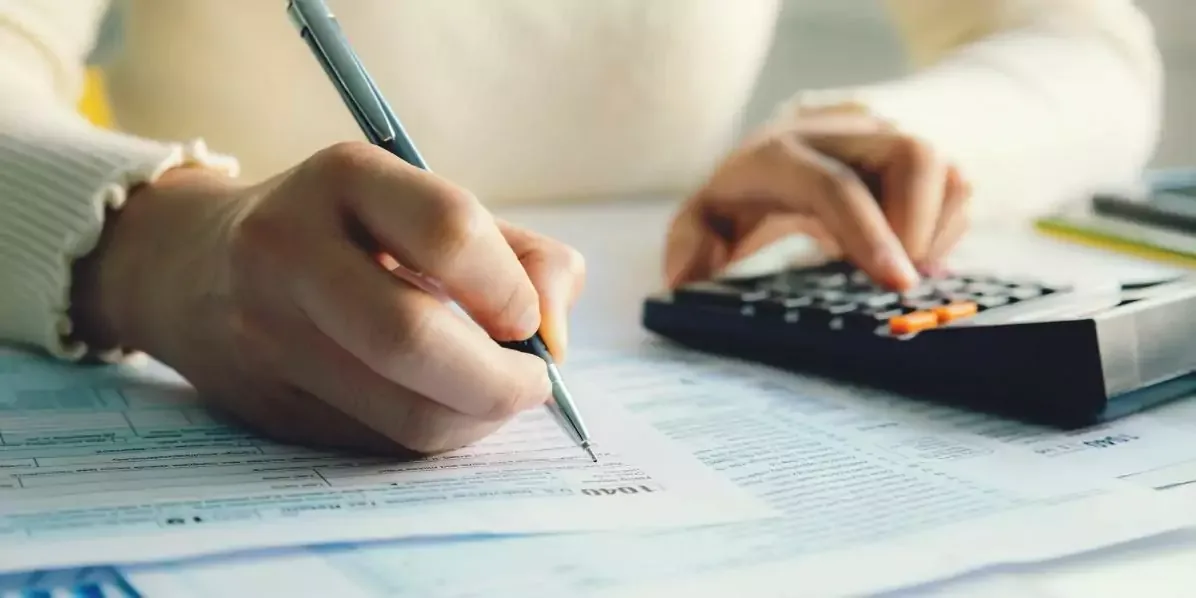 Manos anotando cifras en una planilla impresa mientras usan una calculadora de escritorio.